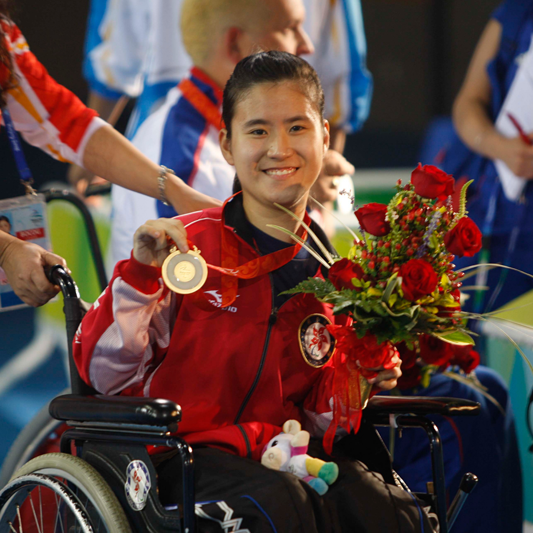 In her wheelchair, Athlete Karen Kwok joyfully celebrates a victory, holding up her gold medal and a bouquet of red roses.