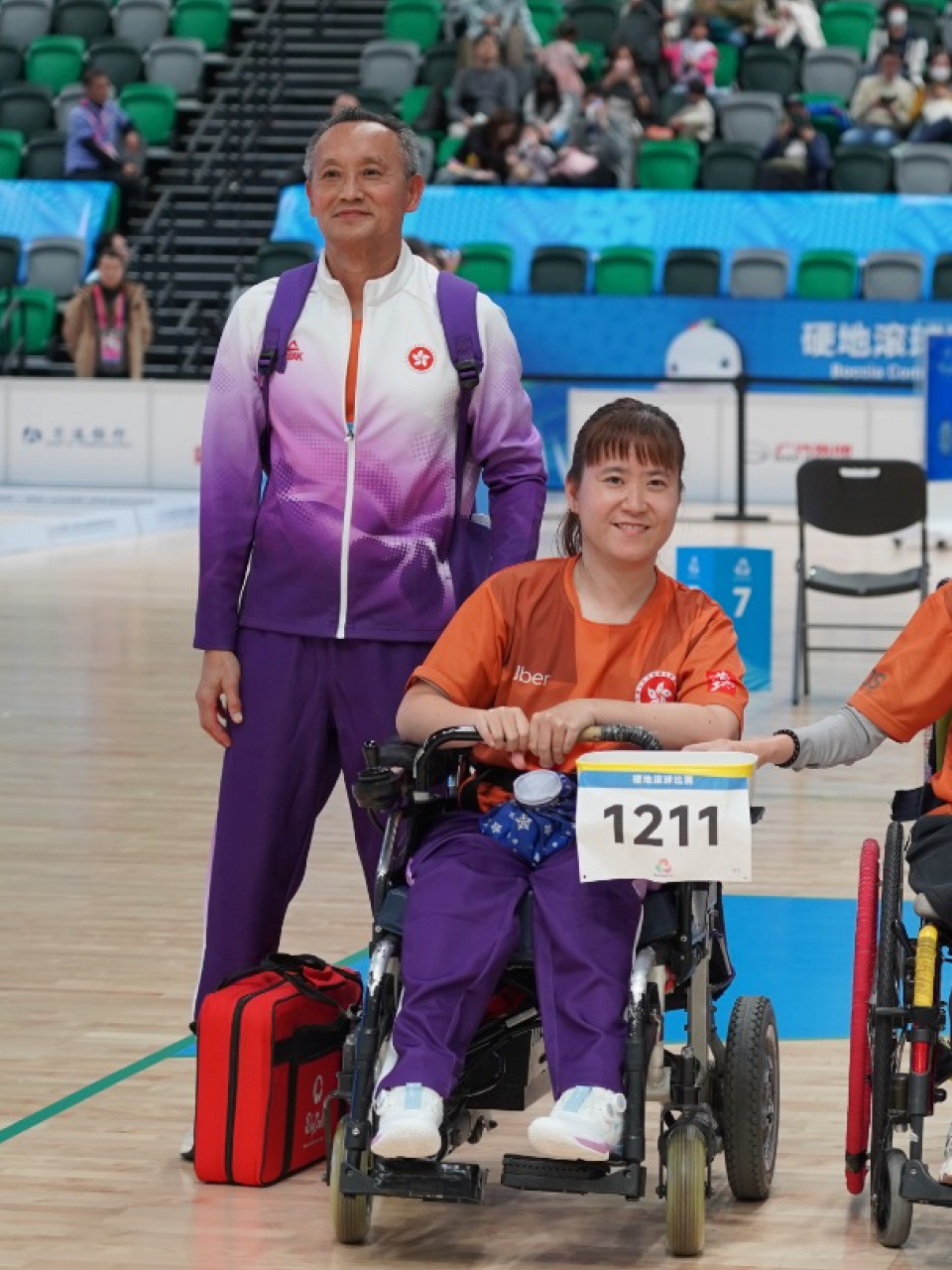 Sharing a proud moment on court, Karen Kwok smiles for a photo in her wheelchair beside her coach in their team uniform.